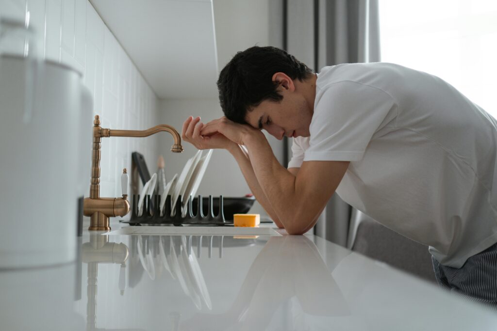 A young man appears tired and stressed, leaning on a kitchen counter in contemplation.