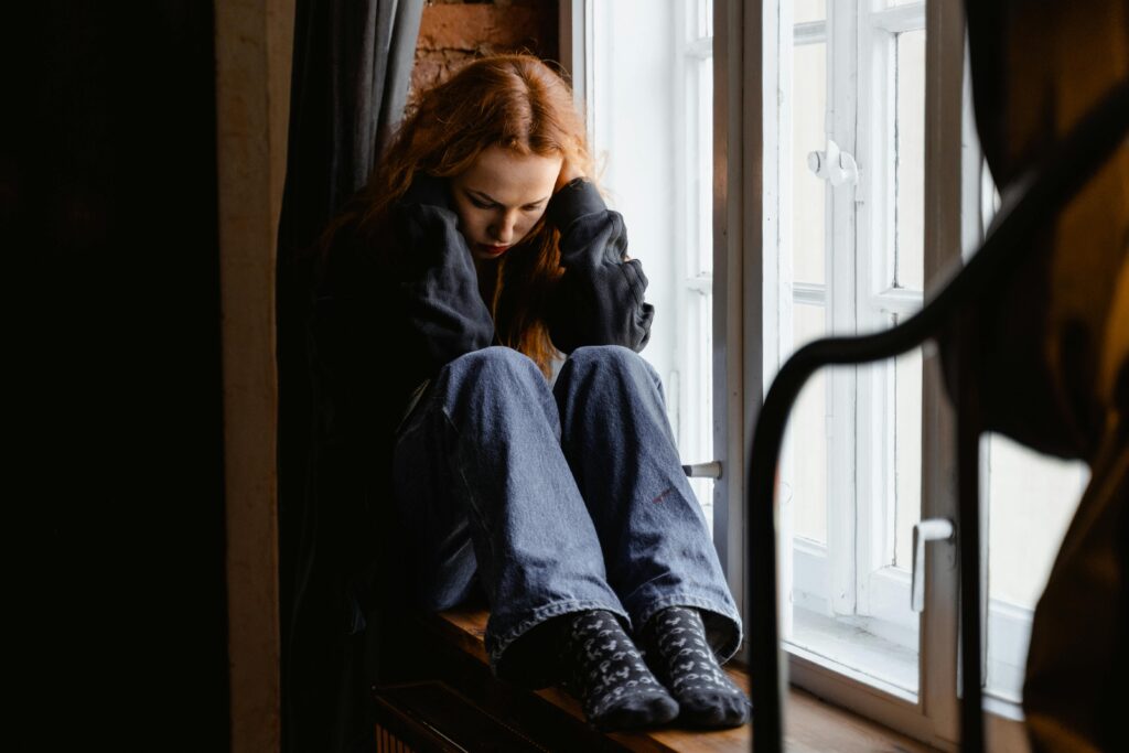 A woman sits pensively by a window, expressing feelings of solitude and contemplation.